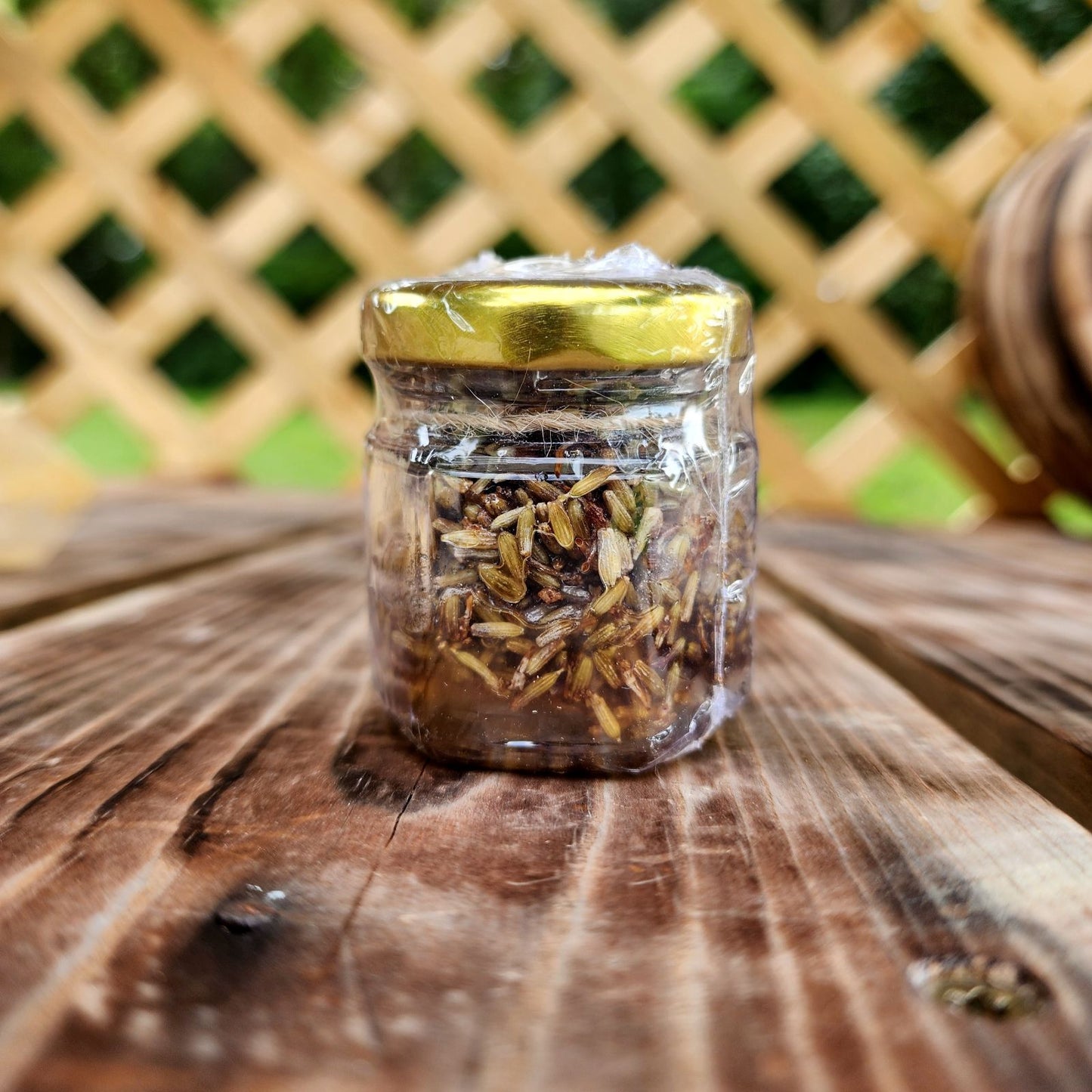 A small glass jar filled with organic lavender-infused honey, placed on a wooden surface. The jar has a golden lid and is tightly sealed, with a blurred green lattice background providing a natural aesthetic.