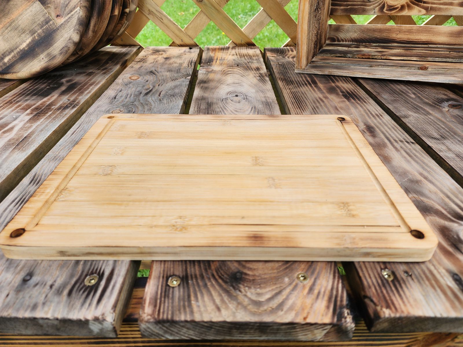 Custom Engraved Cutting Board's A rectangular 9.5 x 13 bamboo cutting board displayed vertically on a rustic wooden table, with a lattice fence and a weathered wooden crate in the background. The cutting board features rounded corners with small holes in each corner for easy hanging or handling. The natural bamboo grain and clean design make it a functional and stylish addition to any kitchen. Custom Engraved Cutting Board's