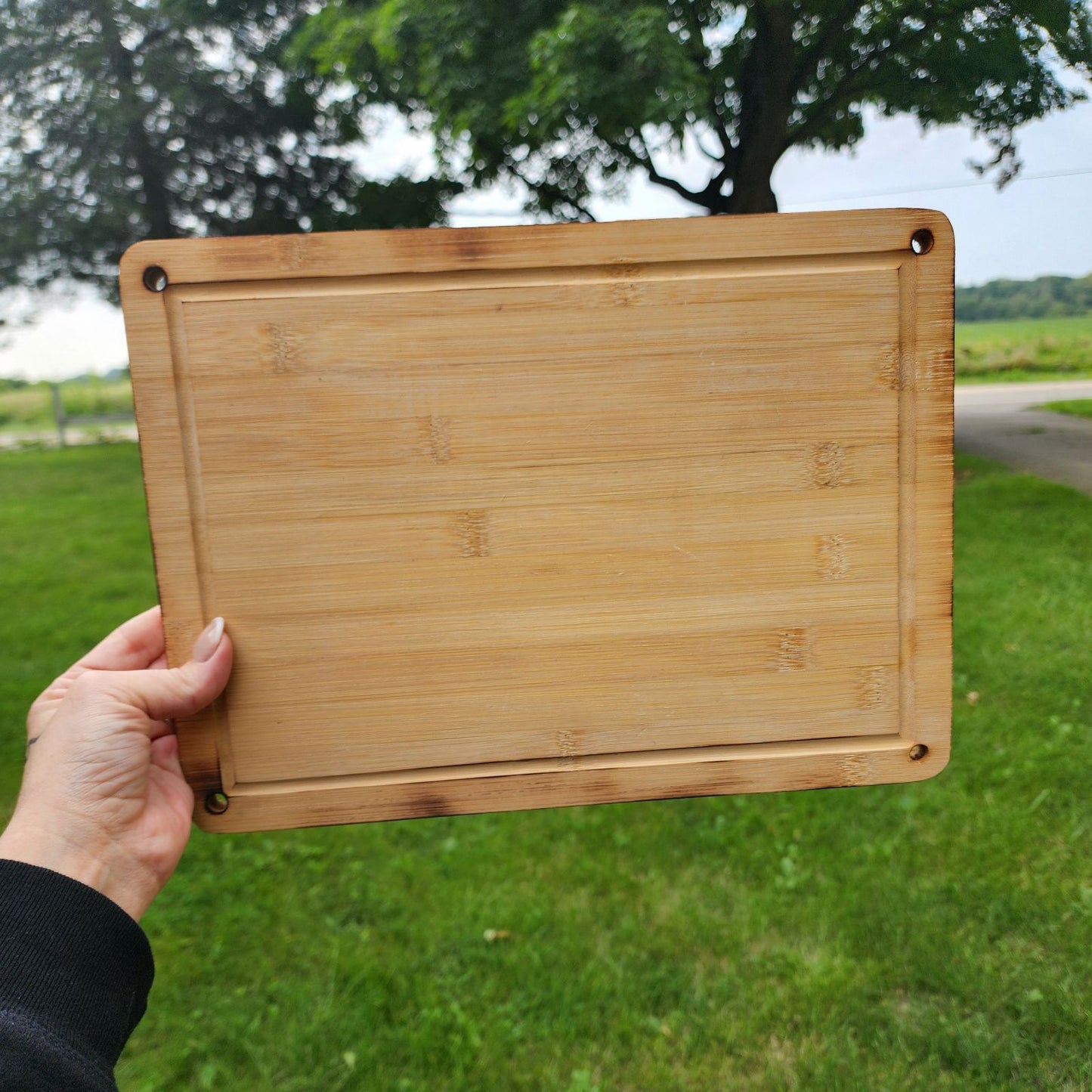 Custom Engraved Cutting Board's A hand holding a rectangular 9.5 x 13 bamboo cutting board with rounded corners and small holes in each corner. The cutting board is displayed outdoors against a backdrop of green grass and a scenic landscape. The natural bamboo grain and smooth finish emphasize its sturdy yet elegant design, perfect for kitchen tasks or serving food. Custom Engraved Cutting Board's