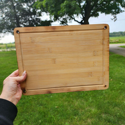 Custom Engraved Cutting Board's A hand holding a rectangular 9.5 x 13 bamboo cutting board with rounded corners and small holes in each corner. The cutting board is displayed outdoors against a backdrop of green grass and a scenic landscape. The natural bamboo grain and smooth finish emphasize its sturdy yet elegant design, perfect for kitchen tasks or serving food. Custom Engraved Cutting Board's