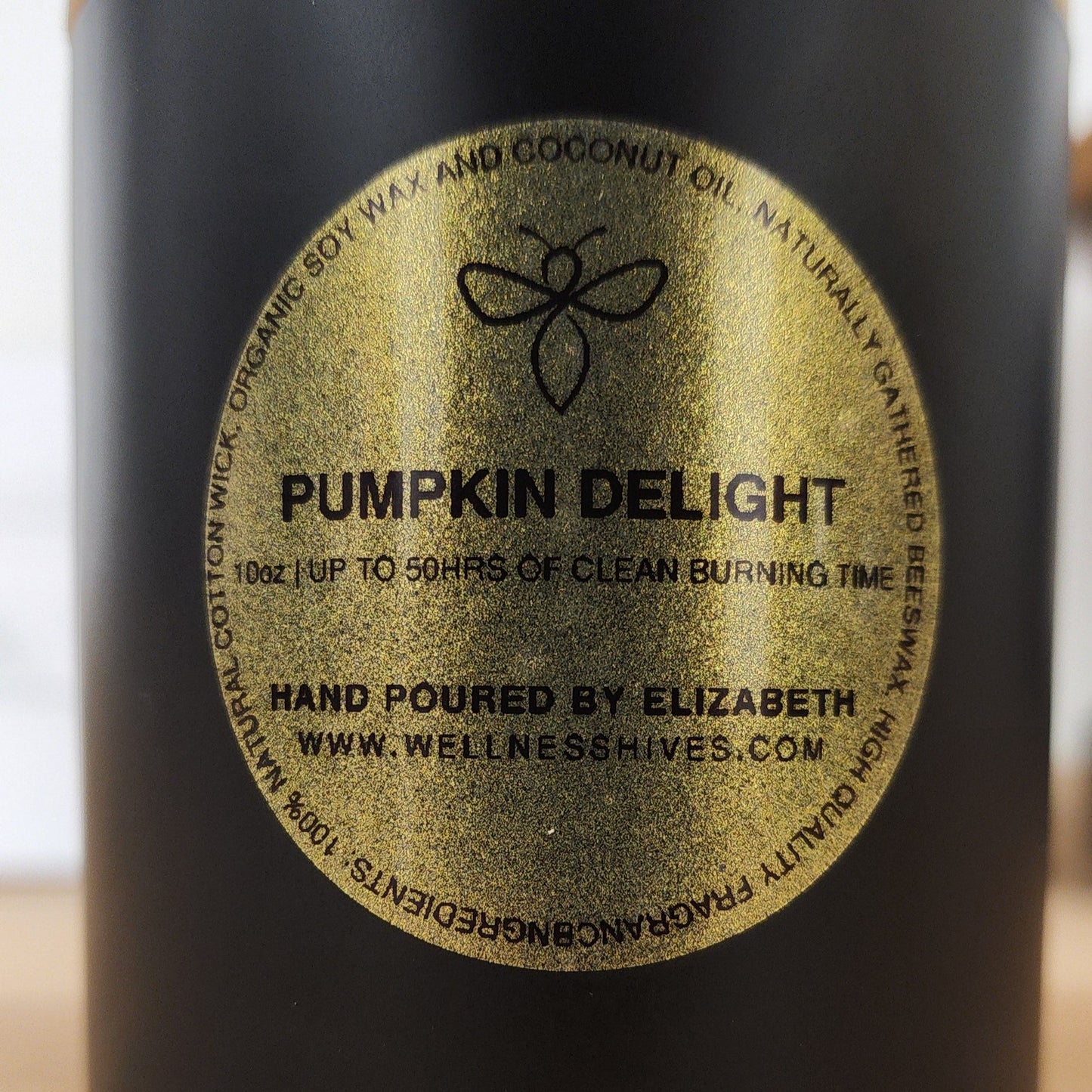 Close-up image of a candle label reading "Pumpkin Delight," showcasing a 10 oz candle with up to 50 hours of clean burning time. The candle is hand-poured by Elizabeth, made from organic soy wax and coconut oil, and is available at www.wellnesshives.com.
