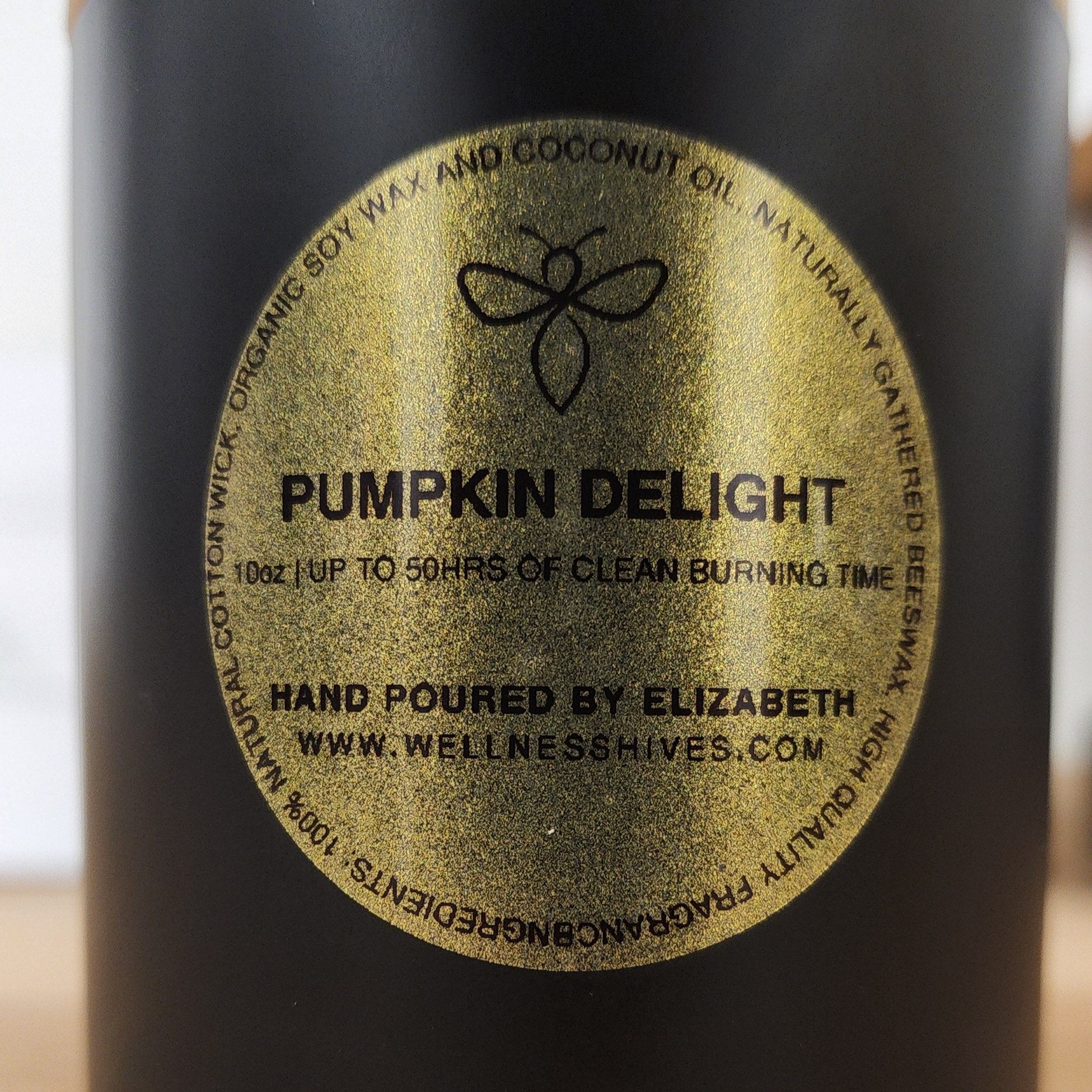 Close-up image of a candle label reading "Pumpkin Delight," showcasing a 10 oz candle with up to 50 hours of clean burning time. The candle is hand-poured by Elizabeth, made from organic soy wax and coconut oil, and is available at www.wellnesshives.com.
