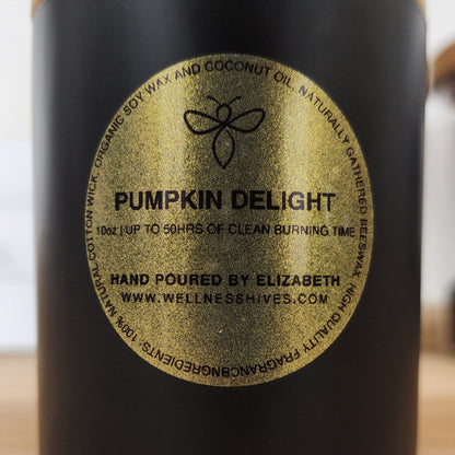 A close-up of the label on a candle jar that reads "Pumpkin Delight," with details of up to 50 hours of clean burning time. The label indicates the candle is hand-poured by Elizabeth and includes a website link: www.wellnesshives.com.