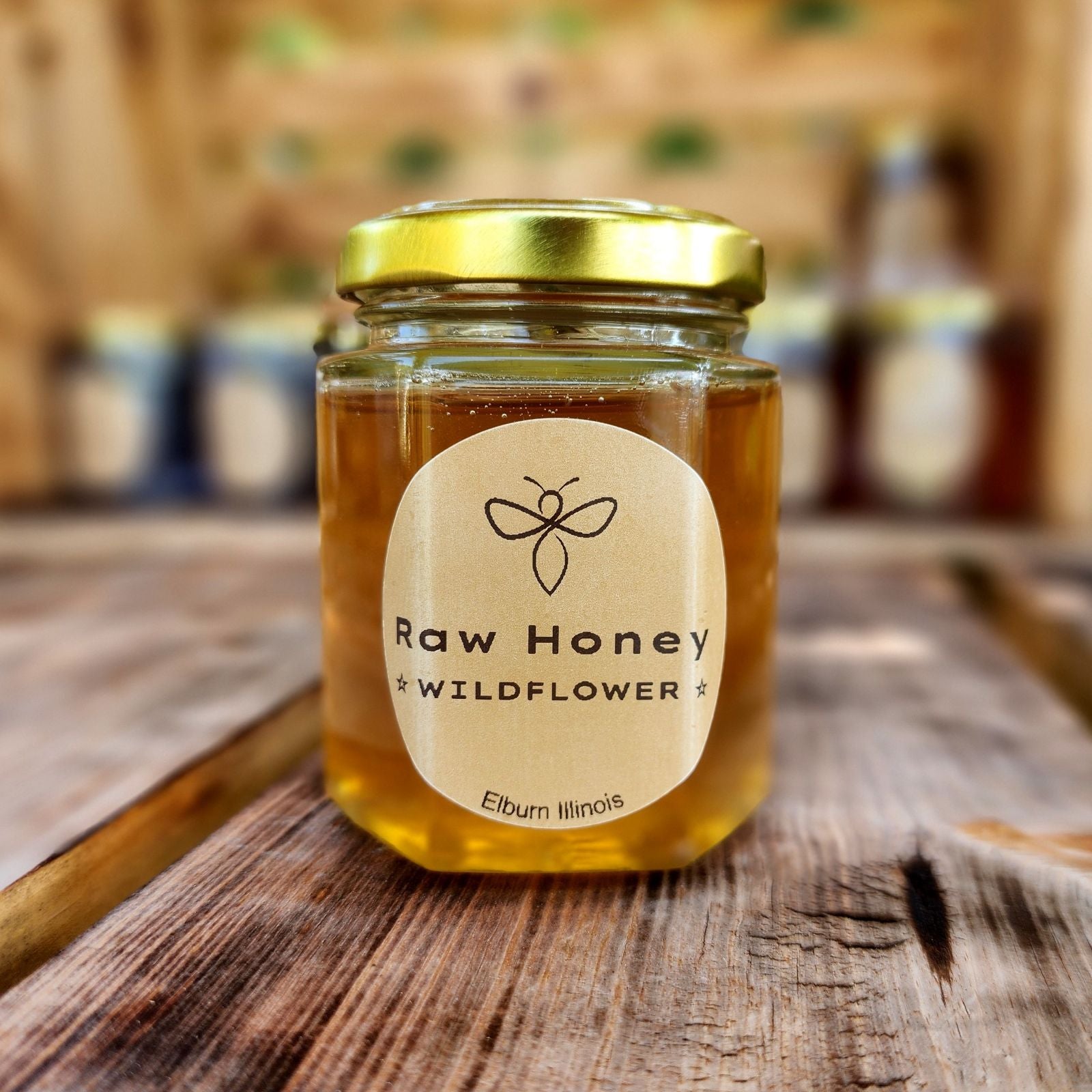 A 10 oz jar of Raw Wildflower Honey sits on a wooden surface with a warm indoor background. The jar has a golden lid and a label that reads 'Raw Honey Wildflower' from Elburn, Illinois. The golden honey inside shines brightly, adding a natural and rustic charm.