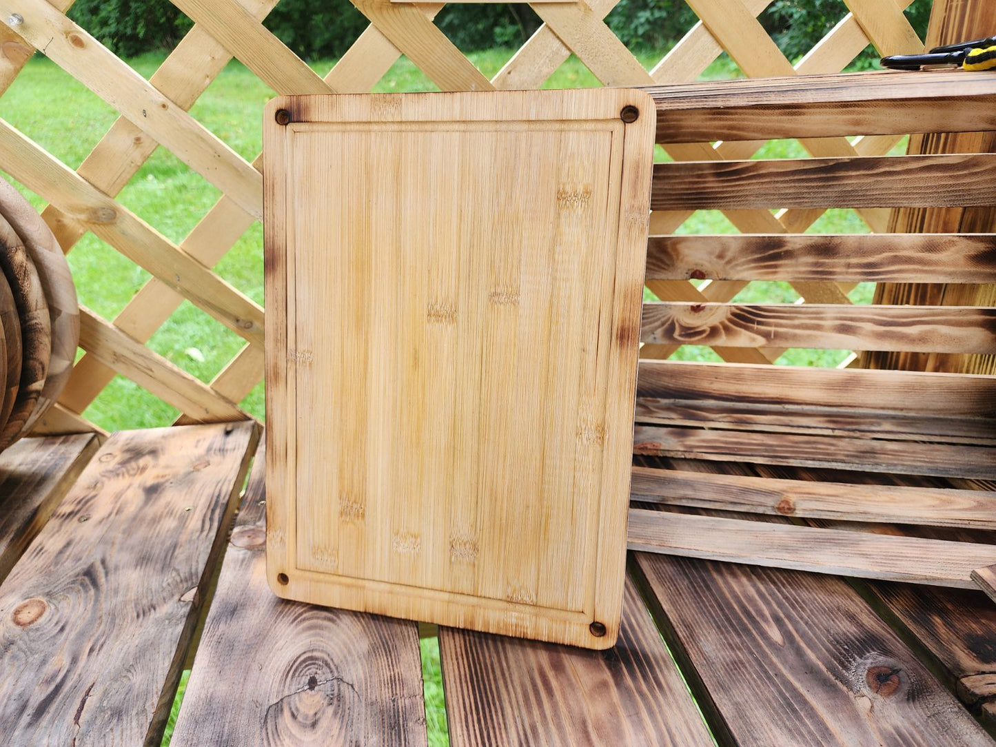 Custom Engraved Cutting Board's A rectangular 9.5 x 13 bamboo cutting board displayed outdoors on a rustic wooden table, with a lattice fence and wooden crate in the background. The cutting board features rounded corners with small holes, adding a touch of design to its simple and functional look. The natural bamboo grain and smooth finish highlight its handcrafted quality, ideal for kitchen use or as a serving board. Custom Engraved Cutting Board's